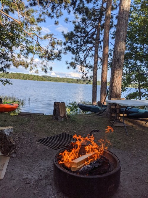 A campfire burns in a fire pit by a serene lake, surrounded by trees and kayaks in the background.