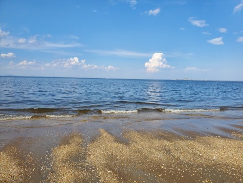 A serene beach scene with gentle waves lapping at the shore under a clear blue sky with fluffy clouds.