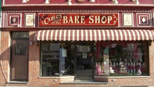 Front view of Carlo's Bake Shop, featuring a red and gold sign, striped awning, and large display windows.