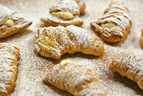 A close-up of freshly baked croissants dusted with powdered sugar, some filled with cream, on a wooden surface.