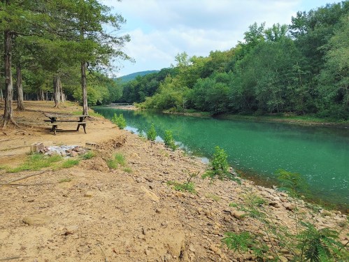 A serene riverbank with green water, surrounded by trees and a picnic table on the shore.