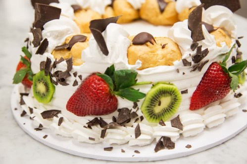 A decorative cake topped with cream, strawberries, kiwi, chocolate shavings, and cream puffs, arranged in a circular shape.