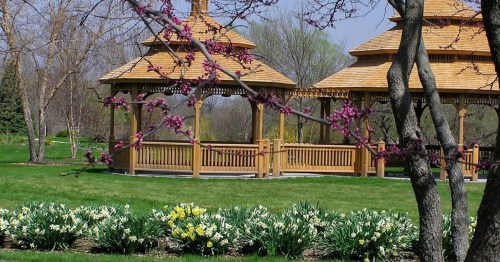 Two wooden gazebos surrounded by blooming flowers and trees in a lush green park.