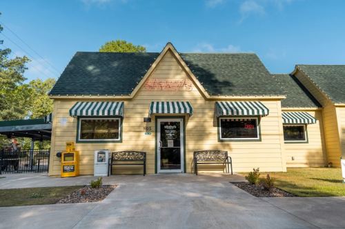 A yellow building with green awnings, benches outside, and a sign indicating it's open. Trees in the background.