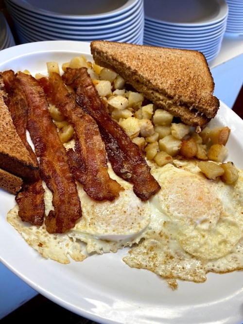 A plate of breakfast featuring two fried eggs, crispy bacon, diced potatoes, and toasted bread.