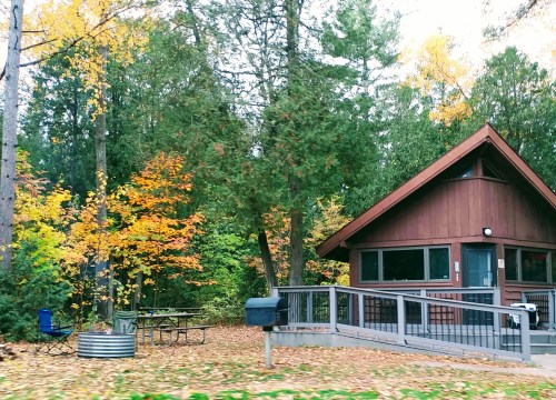 A cozy cabin surrounded by colorful autumn trees, with a picnic table and chairs nearby.