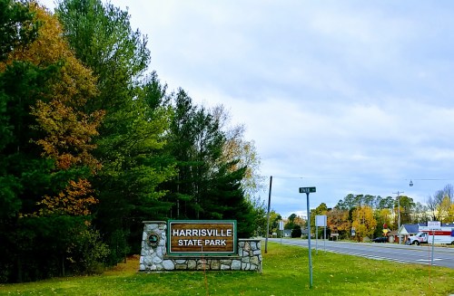 Sign for Harrisville State Park, surrounded by trees and autumn foliage, with a road visible in the background.