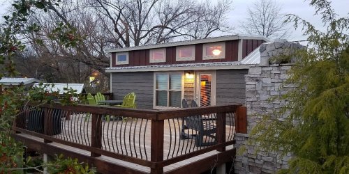 A cozy cabin with a wooden deck, surrounded by trees, featuring colorful chairs and a stone wall.