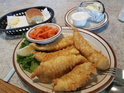 A plate of golden fried fish fillets with lettuce, a lemon wedge, a side of carrots, and a baked potato with sour cream.
