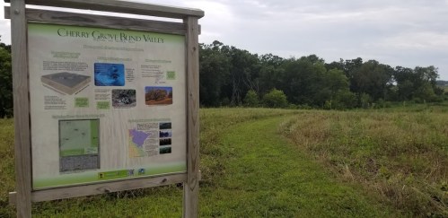 Sign for Cherry Grove Blind Valley with informational graphics, surrounded by green grass and trees under a cloudy sky.