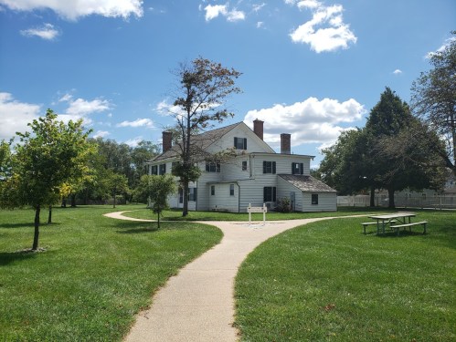 A white house with a sloped roof surrounded by green grass and trees, with a pathway leading to it under a blue sky.