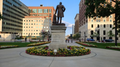Statue of a man in a suit on a pedestal, surrounded by colorful flower beds in an urban park setting.
