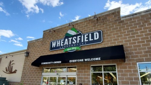 Sign for Wheatsfield Cooperative, featuring "Community-Owned" and "Everyone Welcome," against a blue sky.