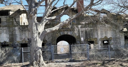 An abandoned, crumbling building with a large archway, surrounded by a chain-link fence and a bare tree.