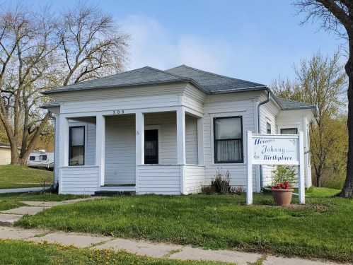 White house with a gray roof, featuring a sign that reads "Herbert Johnny Cash Birthplace" and a small flower bed.