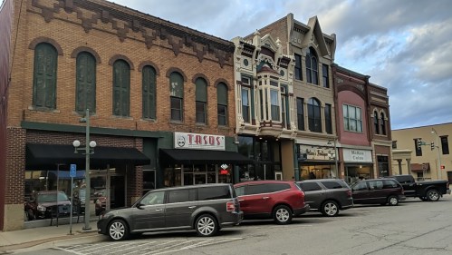 Historic buildings line a street, featuring a taco restaurant and a mix of shops, with parked cars in front.