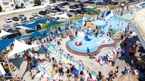 A busy water park scene with families enjoying pools, slides, and sun loungers on a sunny day.