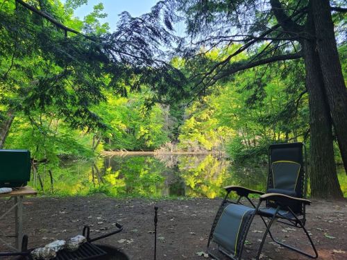 A serene campsite by a calm lake, surrounded by lush green trees and reflections on the water. Two chairs are set up nearby.