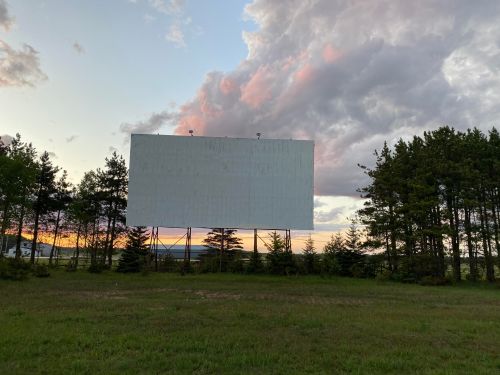 A blank outdoor movie screen stands among trees, with a colorful sky and clouds in the background at dusk.