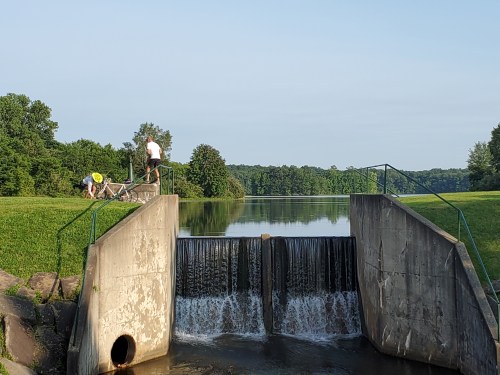 Two people work near a small dam, with water flowing over it and a serene landscape in the background.
