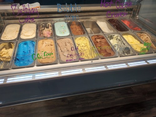 A display of various ice cream flavors in metal tubs, labeled with colorful names like Birthday Cake and Hot Cocoa Crunch.