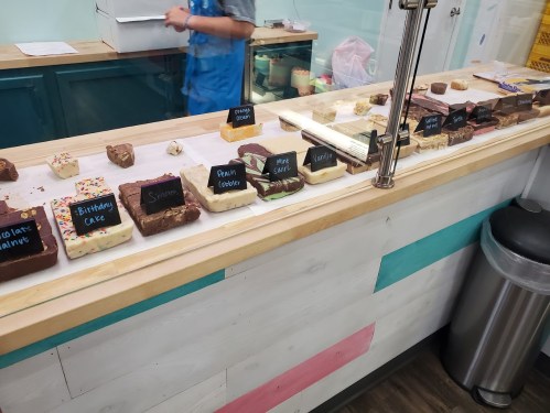 A display of various fudge flavors on a counter, with labels for each type, in a colorful shop setting.
