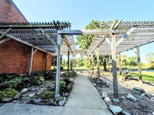 A pathway leads through a wooden trellis structure, surrounded by greenery and rocks, under a clear blue sky.