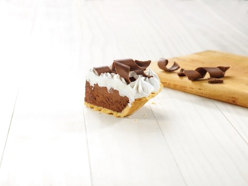 A slice of chocolate cream pie topped with whipped cream and chocolate shavings on a wooden surface.