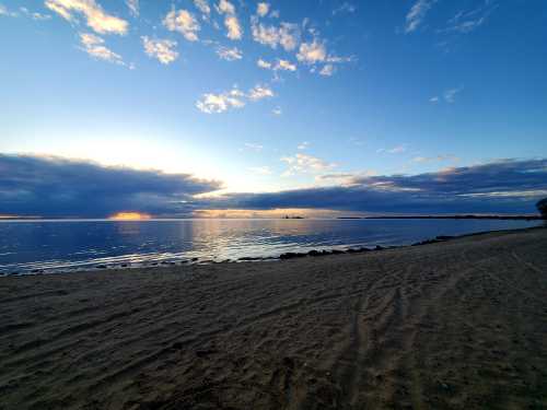 A serene beach at sunset, with calm waters reflecting the sky and clouds, and a sandy shore in the foreground.