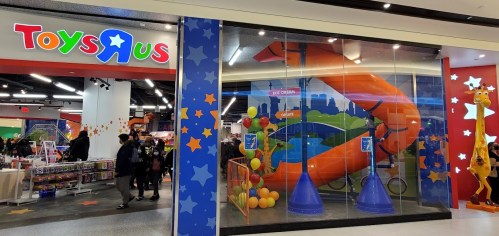 Toys "R" Us storefront featuring colorful decorations and a large inflatable slide inside. Shoppers visible outside.