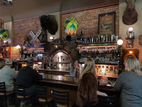 A rustic bar with a brick wall, shelves of liquor, and patrons seated at the counter under colorful pendant lights.