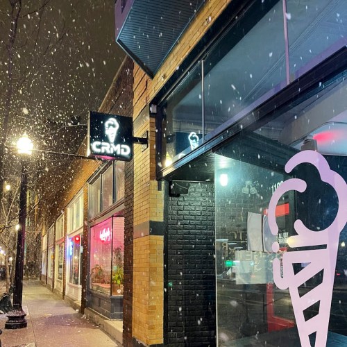 A snowy night scene outside an ice cream shop with a neon sign, showcasing a cozy urban atmosphere.