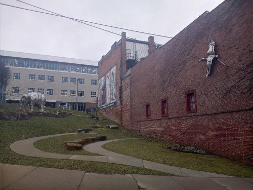 A brick wall with a mural, a sculpture of a horse, and a curved pathway in a grassy area near a building.