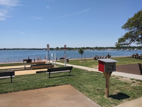 A sunny waterfront park with benches, a sandy area, and a view of boats on the water.