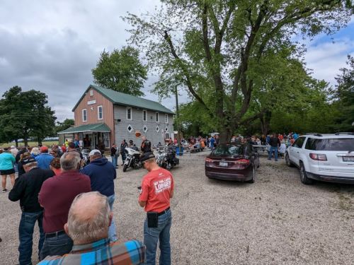 A large crowd gathers outside a building with a green roof, surrounded by trees and parked cars.