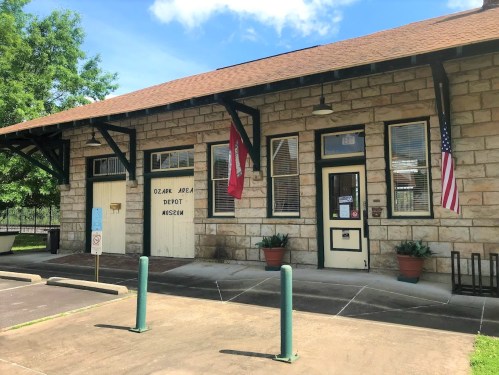 Historic stone building with flags, labeled "Ozark Area Depot Museum," surrounded by greenery and a paved area.