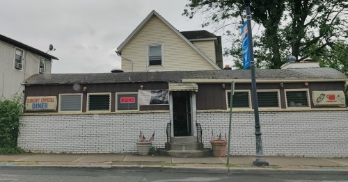 A small diner with a white brick exterior and a sign, set against a cloudy sky and surrounded by greenery.