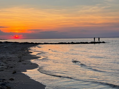 Two people stand on rocks by the shore at sunset, with vibrant colors reflecting on the calm water.