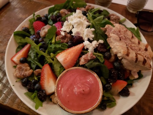 A colorful salad with mixed greens, strawberries, blueberries, grilled chicken, goat cheese, and a pink dressing.