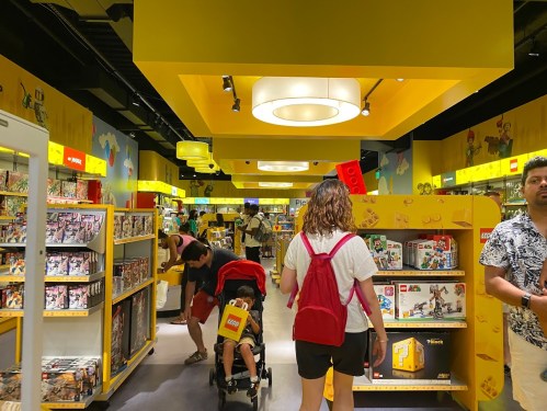 A vibrant toy store filled with colorful shelves, shoppers, and a child in a stroller, under bright yellow lighting.