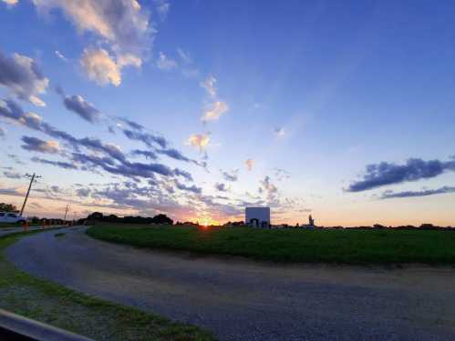 A scenic sunset over a field, with colorful clouds and rays of light illuminating the sky. A dirt road leads into the scene.