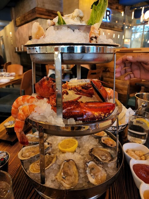 A seafood tower featuring lobster, shrimp, oysters, and crab on ice, garnished with lemon and lettuce.