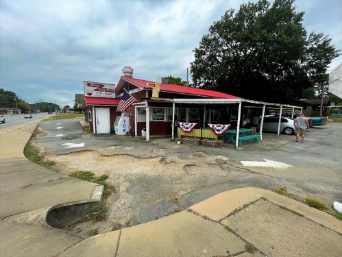 A small roadside shop with a red roof, patriotic decorations, and a sign for ice, surrounded by a gravel parking area.