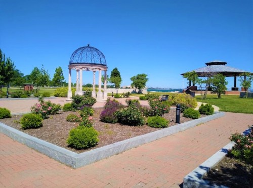 A sunny park scene featuring a gazebo, flower beds, and a view of the water in the background.