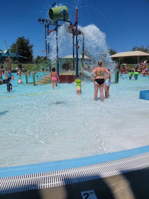 A water play area with children and adults enjoying splashes from a large water feature on a sunny day.