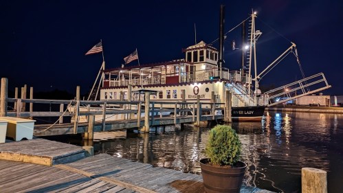 A riverboat docked at night, illuminated with lights, surrounded by water and wooden walkways.