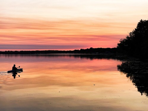 A person kayaking on a calm lake at sunset, with vibrant pink and orange hues reflecting on the water.
