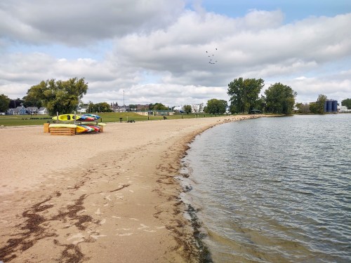 A sandy beach with colorful kayaks on the shore, surrounded by green trees and a calm water body under a cloudy sky.