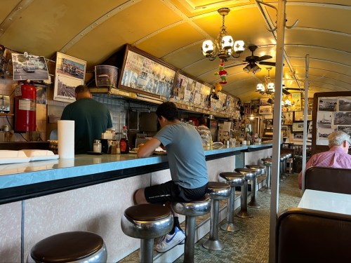 A retro diner interior with a long counter, stools, and vintage photos on the walls. Two patrons are seated.