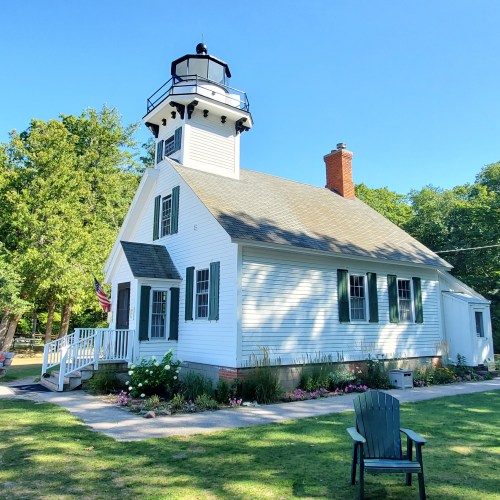 A white lighthouse with a black lantern room, surrounded by greenery and a chair in the foreground.
